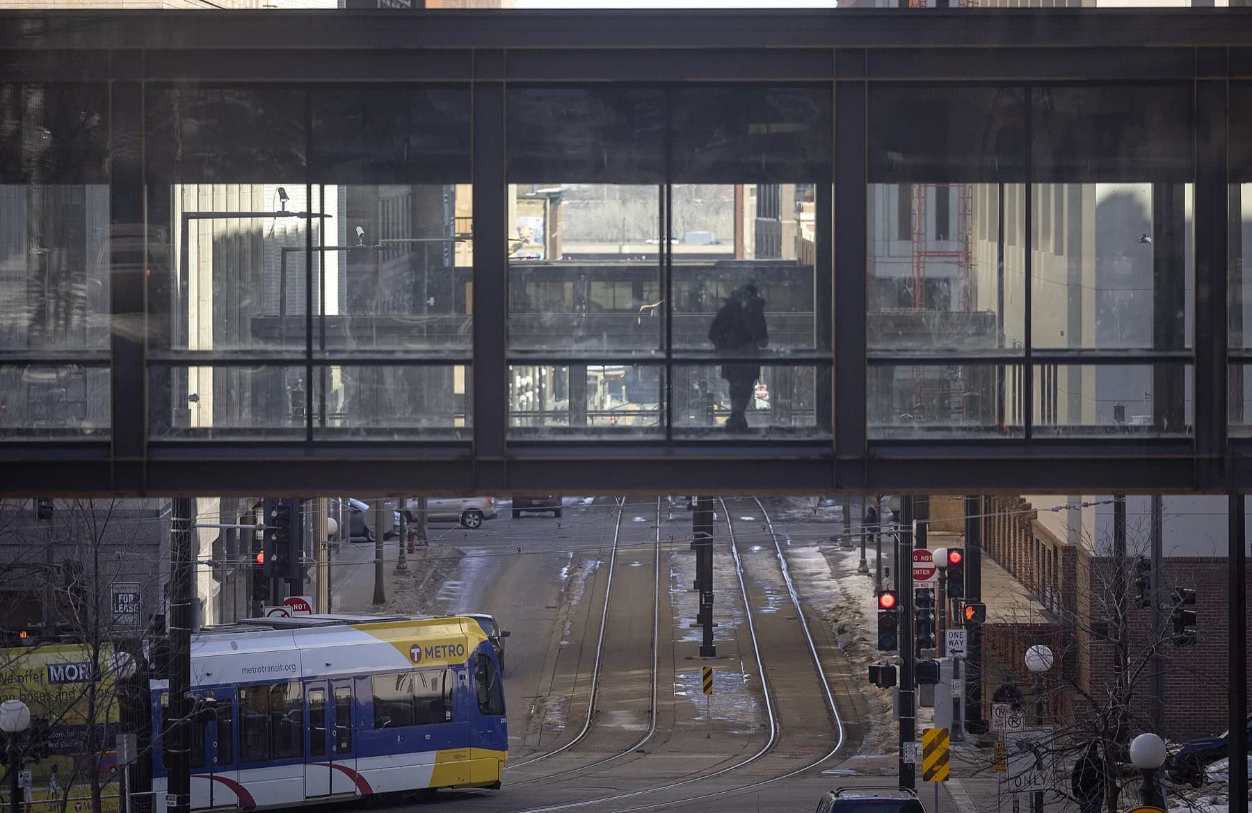 A lone figure walks through a glass skyway at dusk on a winter evening, with a Metro Transit Green Line car passing below on Cedar Street.